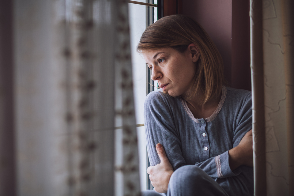 Mujer sentada mirando por la ventana cruzando los brazos alrededor de sí misma.