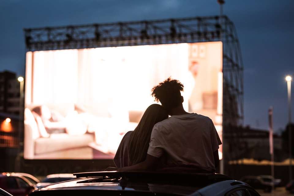 Silhouette of backs of two people sitting with arms around each other, watching an outdoor movie.