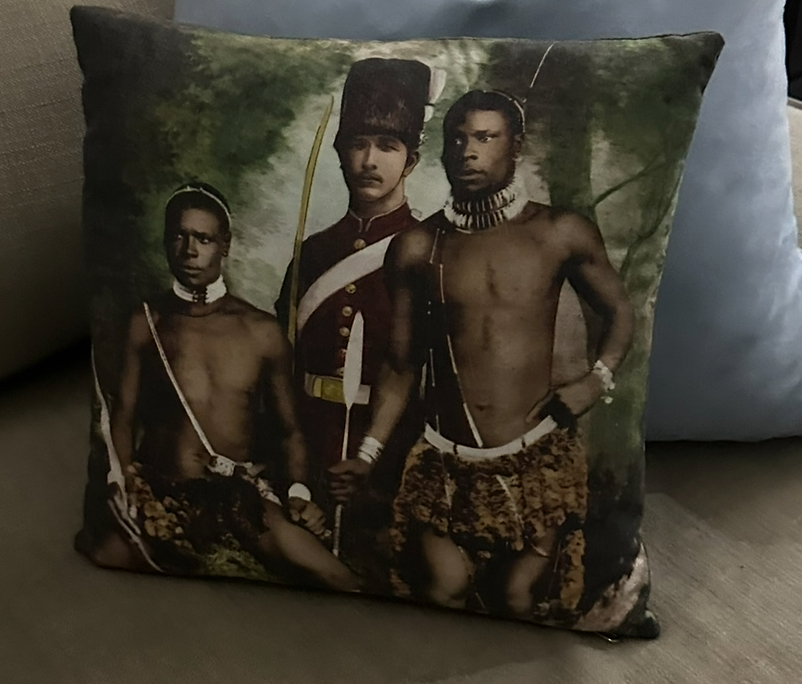 Pillow depicting white man in uniform and two Black men in traditional clothing on bottom and necklaces.