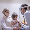 A masked health care professional using a stethoscope to listen to a toddler's chest who is sitting on her mother's lap.