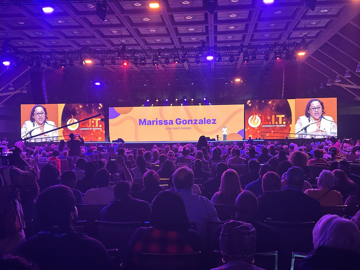 Marissa Gonzalez on a stage with screens behind her showing her - and an audience in front.