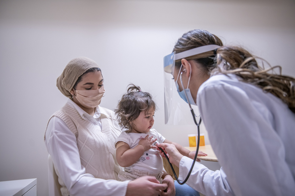 A masked health care professional using a stethoscope to listen to a toddler's chest who is sitting on her mother's lap.