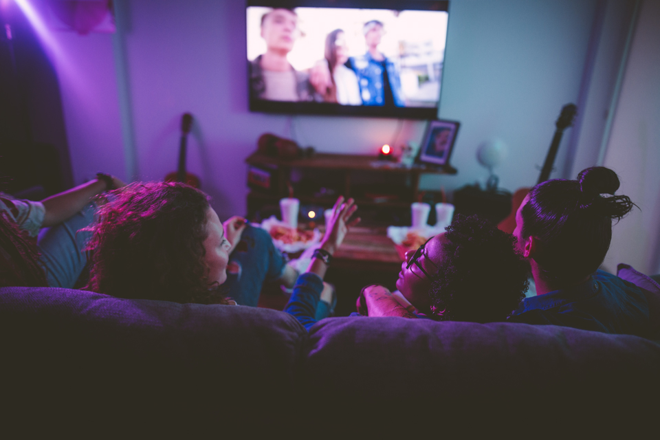 Three teens sitting down, talking and watching TV together at home.