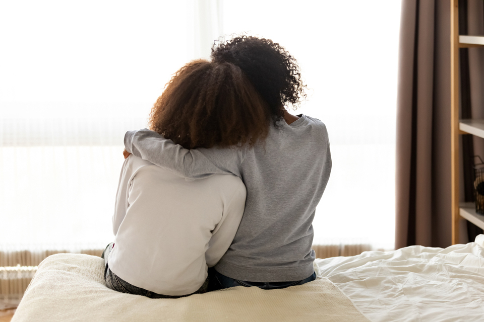 Back view of two people sitting on edge of bed, one with arm around other who leans on her shoulder.