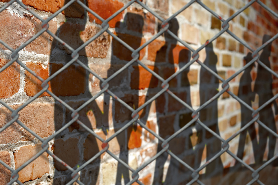 Shadows of people walking in line connected at hands seen on brick wall behind a chain-link fence.