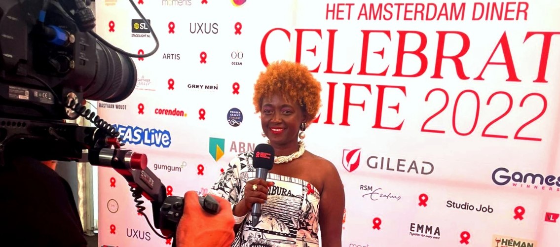 HIVstigmafighter standing in front of Celebrate Life 2022 banner, holding mic and being filmed.