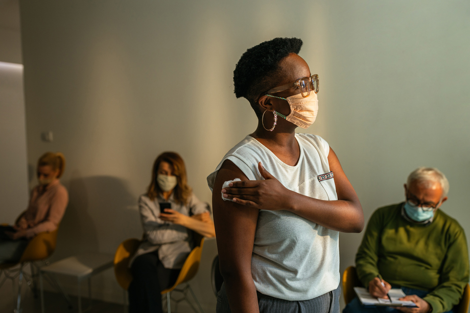 Woman holding an absorbent cotton to her arm after vaccination in a waiting room with other people seated in the background.