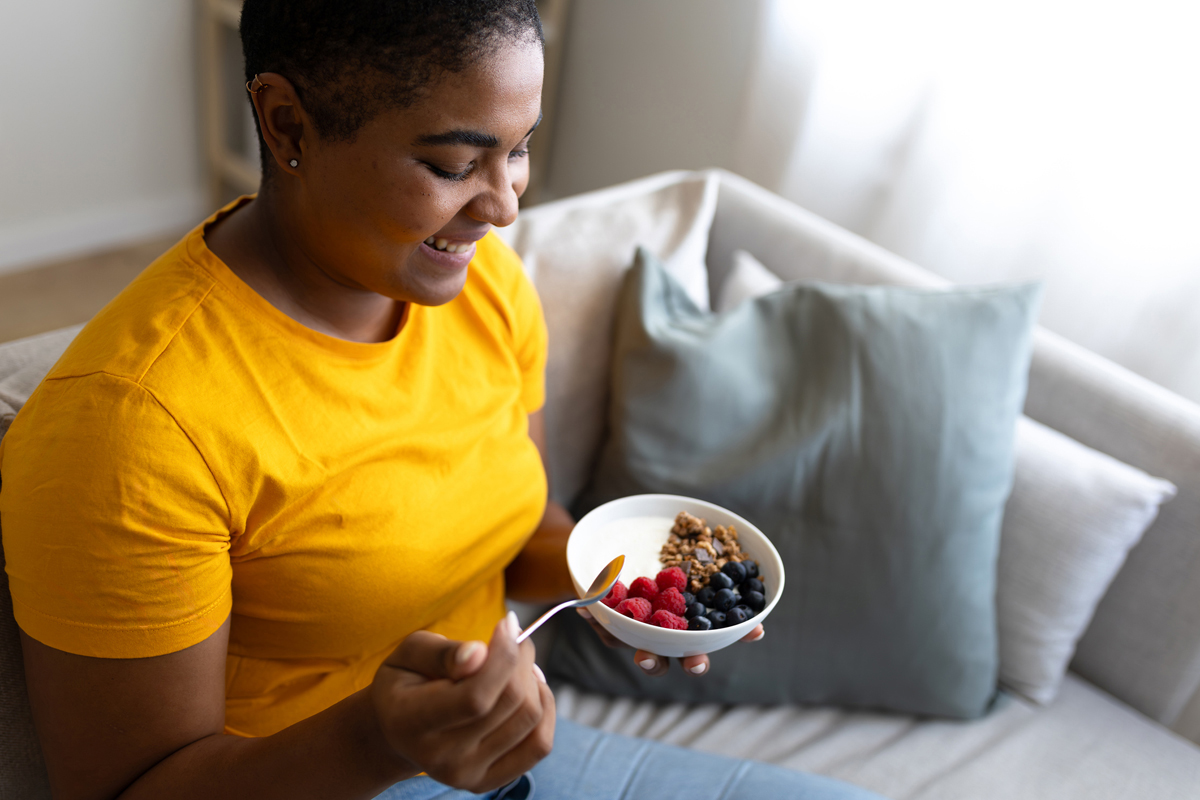 Una persona con una camiseta amarilla, sonriendo, sentada en un sofá, sosteniendo una cuchara y un tazón con yogur, granola y frutos del bosque.