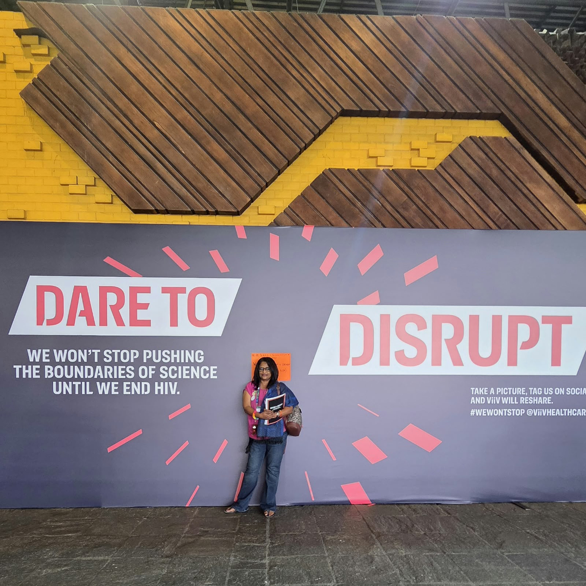 The Well Project global ambassador Jyoti Dhawale standing in front of sign at AIDS 2026 that reads "Dare to Disrupt".