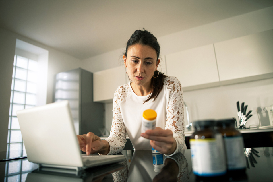 Una mujer frente a una computadora portátil mirando una botella de medicamento.