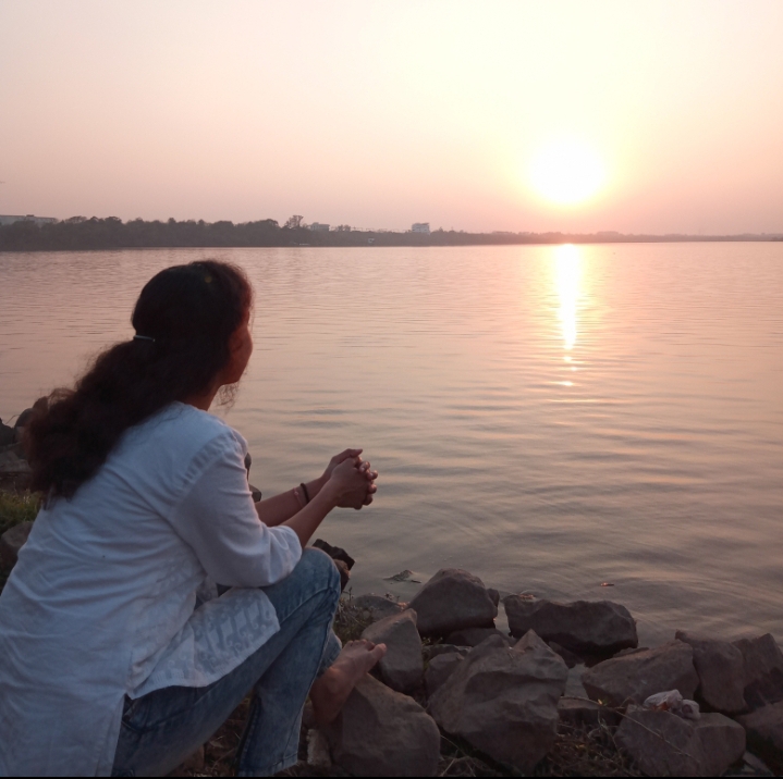 Back view of woman sitting on rocks looking out at water during sunset. 