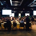 Many people sitting and standing at conference, with multiple screens hanging from ceiling. 