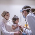A masked health care professional using a stethoscope to listen to a toddler's chest who is sitting on her mother's lap.