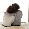 Back view of two people sitting on edge of bed, one with arm around other who leans on her shoulder.