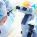 Lab technician in mask, near a microscope, looking at a flask of liquid. 