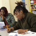 Two people sitting at a desk with binders, textbooks and papers, one looking at papers, one writing.