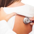 Back of woman in hospital gown; a doctor's hand holds a stethoscope on her back.