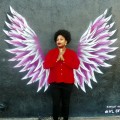 Porchia Dees, standing with palms together, in front of painting of wings on wall by Keenan Chapman.
