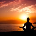 Silhouette of back of person in seated yoga pose near sea at sunset.