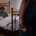 Person on bed and another person standing near bed holding a condom.