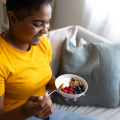 Una persona con una camiseta amarilla, sonriendo, sentada en un sofá, sosteniendo una cuchara y un tazón con yogur, granola y frutos del bosque.