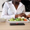 Woman sitting at table with a plate of vegeteables and a bowl in front of her and holding and looking at blood sugar monitor.