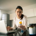 A woman at a laptop looking at medication bottle.