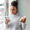 Woman standing near window holding two medication models and looking at phone.