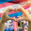 Close up of hands creating shape of heart with people and Trans flags in the background.