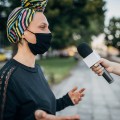 Profile of person in headwrap and mask with a microphone being held to her face by someone's hand..