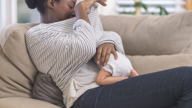 Woman sitting on couch looking under her shirt at infant underneath breastfeeding.