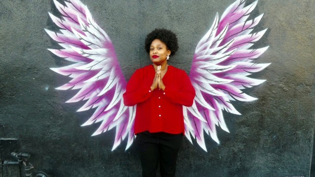 Porchia Dees, standing with palms together, in front of painting of wings on wall by Keenan Chapman.