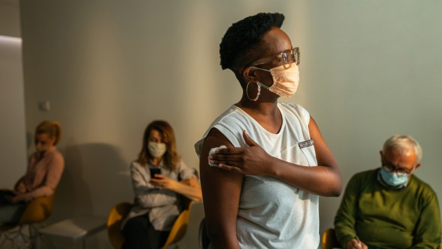 Woman holding an absorbent cotton to her arm after vaccination in a waiting room with other people seated in the background.