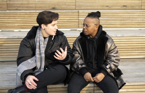 Two people sitting next to each other, outside, having a conversation.