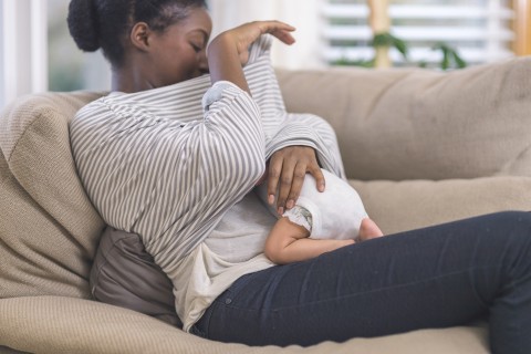 Woman sitting on couch looking under her shirt at infant underneath breastfeeding.