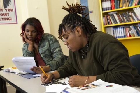 Two people sitting at a desk with binders, textbooks and papers, one looking at papers, one writing.