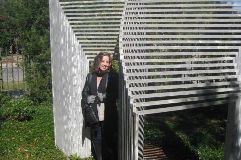 Vickie Lynn, smiling, standing outside in front of abstract metal sculpture in garden.