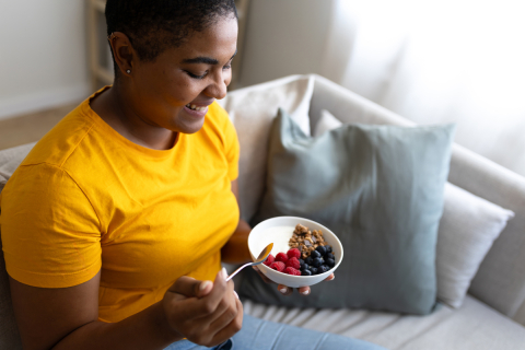 Person in yellow shirt, smiling, sitting on a sofa holding a spoon and a bowl of yogurt, granola, and berries from a bowl.