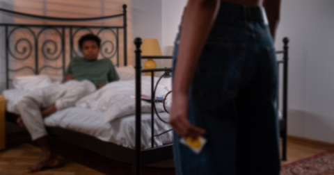 Person on bed and another person standing near bed holding a condom.