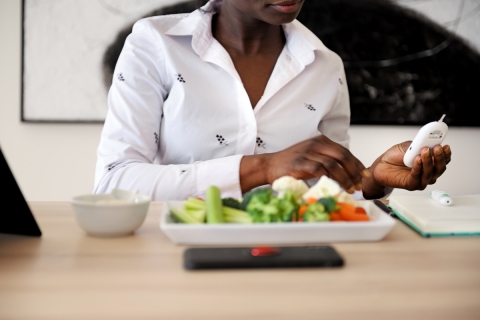 Woman sitting at table with a plate of vegeteables and a bowl in front of her and holding and looking at blood sugar monitor.