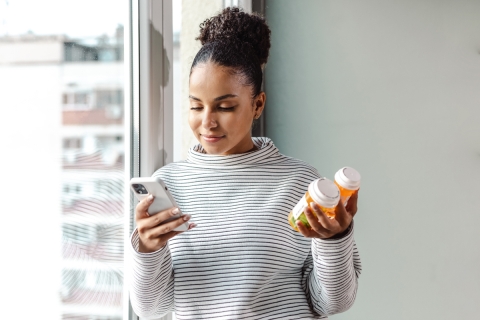 Mujer parada cerca de una ventana sosteniendo dos modelos de medicamentos y mirando el teléfono.