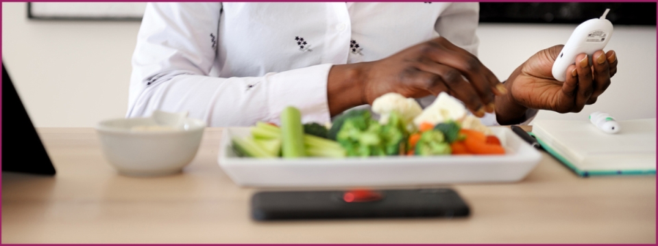 Woman sitting at table with a plate of vegetables and a bowl in front of her and holding and looking at blood sugar monitor.