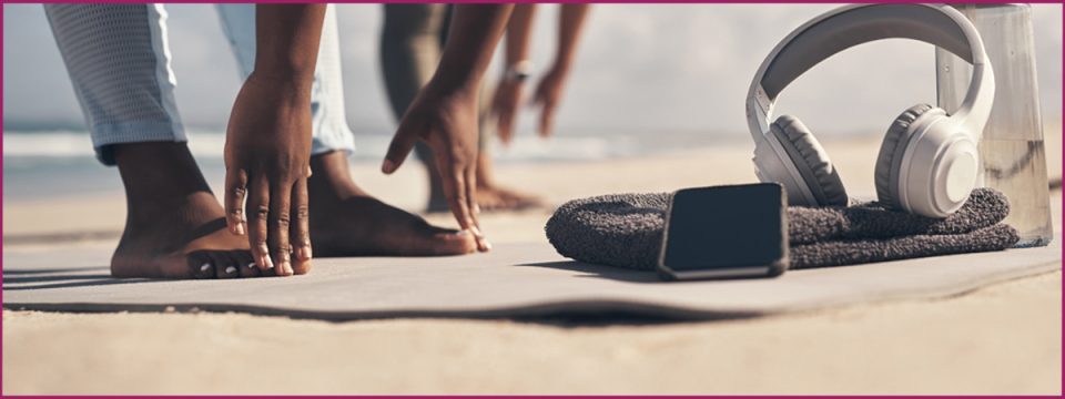 Two women stretching at the beach next to headphones and water bottle.