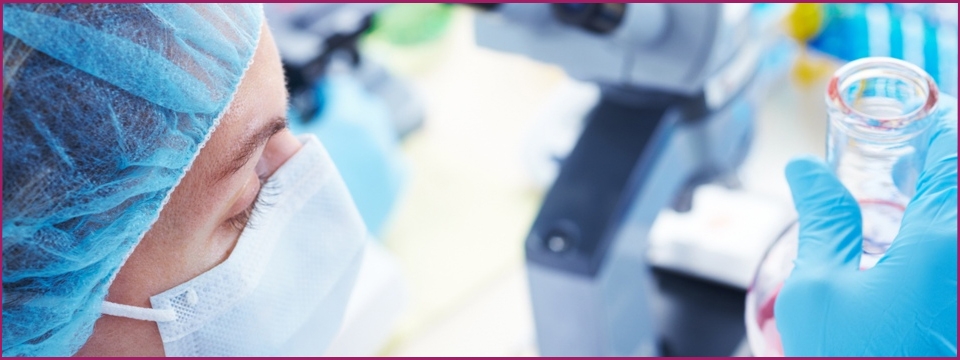 Lab technician in mask, near a microscope, looking at a flask of liquid.