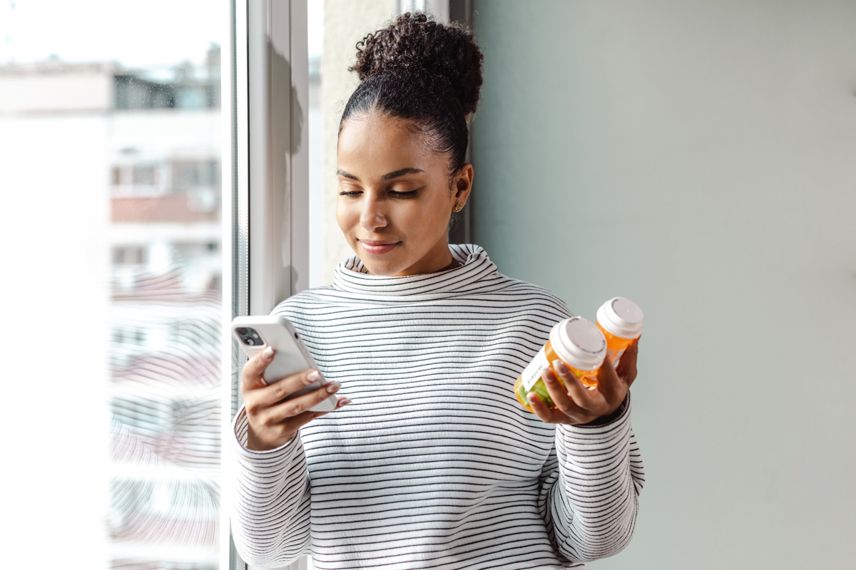 Woman standing near window holding two medication models and looking at phone.